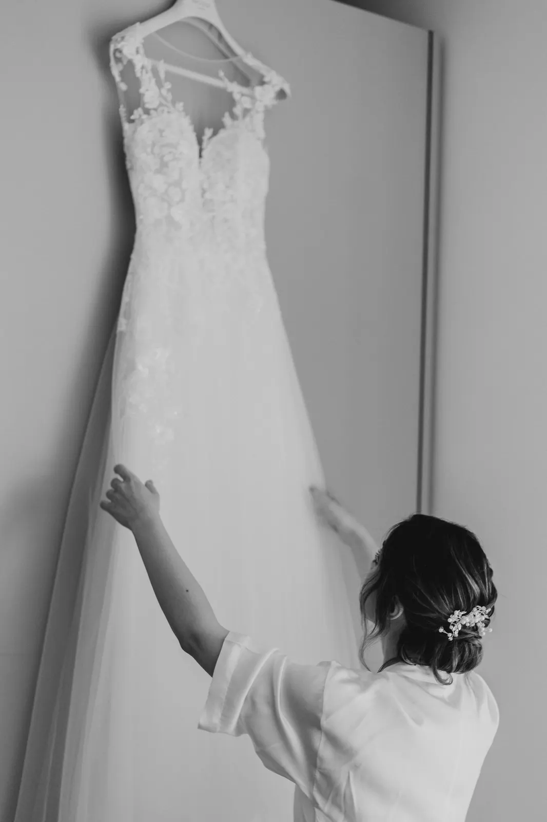 La sposa in vestaglia sistema l'abito in pizzo appeso alla parete, in bianco e nero
