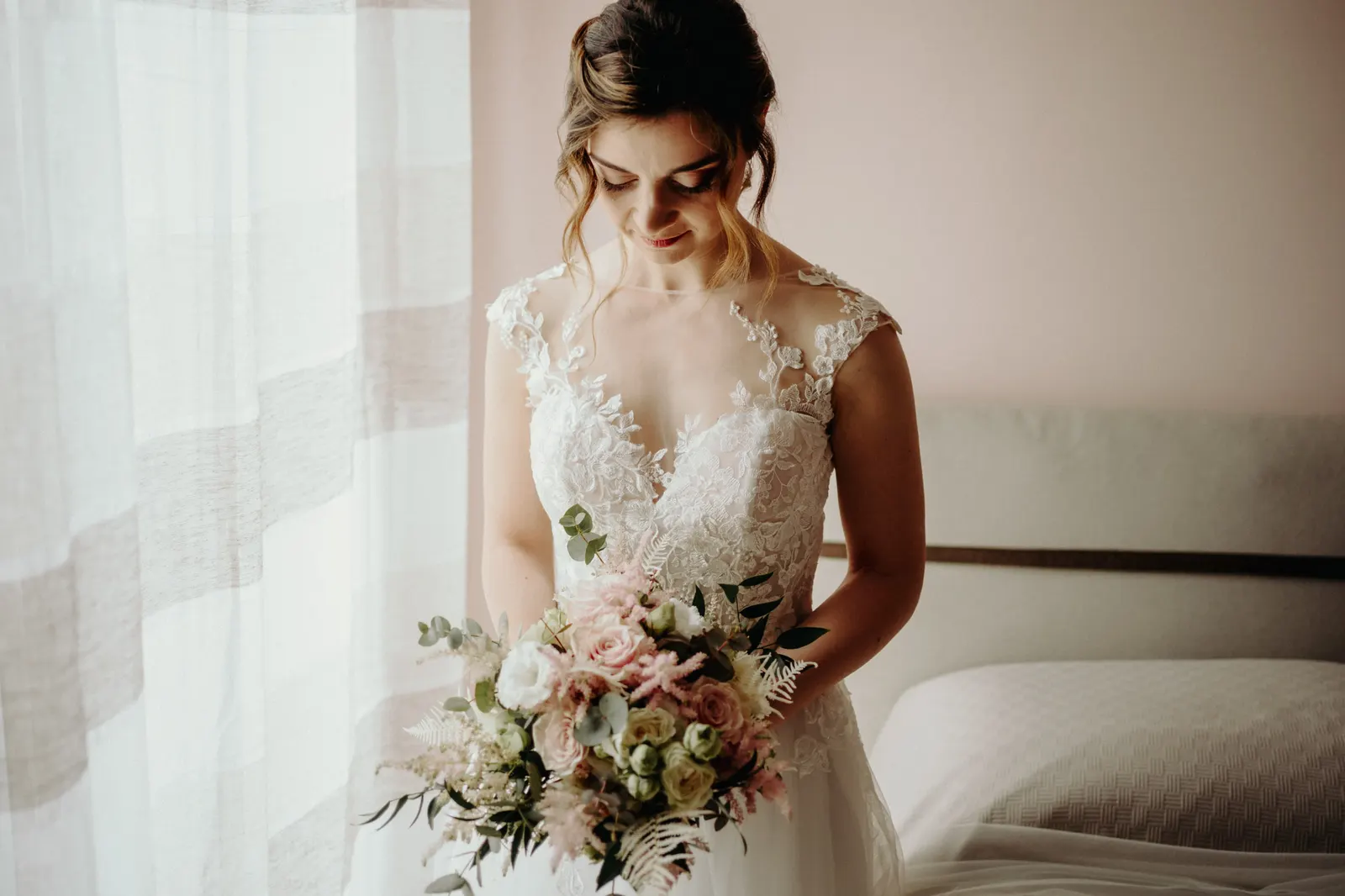La sposa guarda il bouquet accanto alla finestra, nella luce naturale della camera