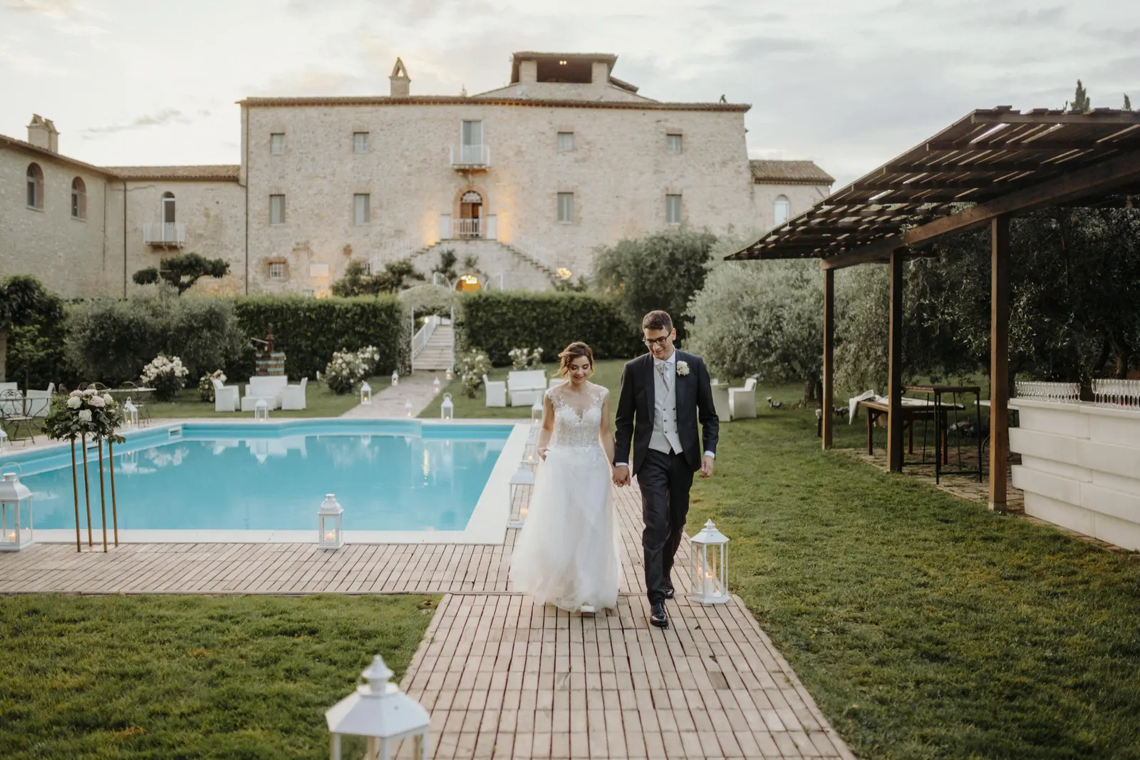 Gli sposi camminano mano nella mano accanto alla piscina, con il Castello di Montignano alle spalle