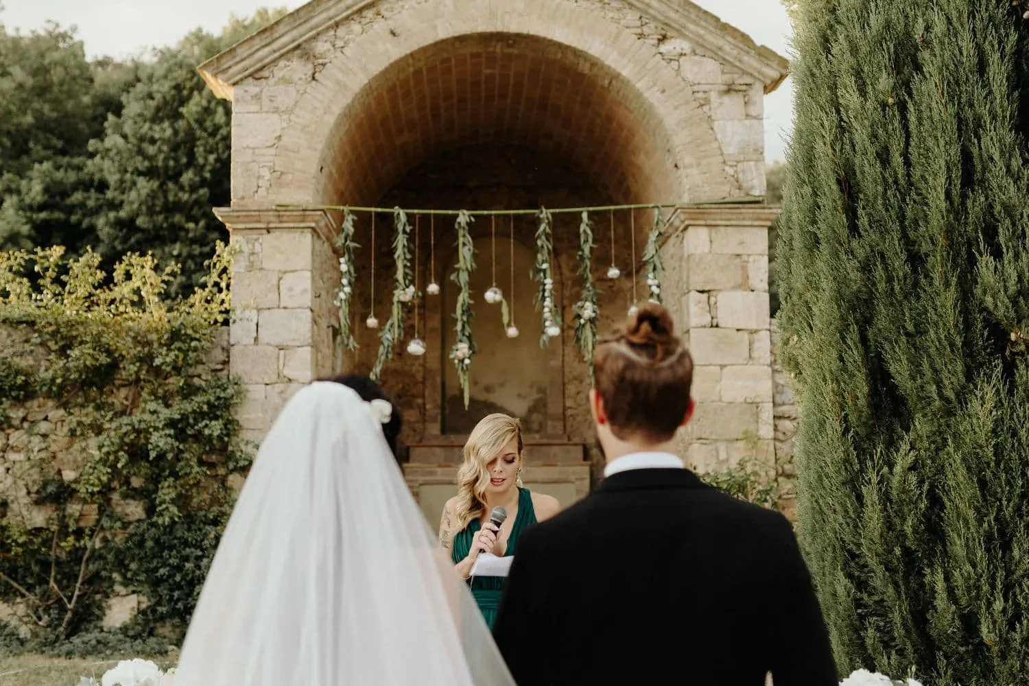 The couple seen from behind in front of the stone shrine with hanging flowers, during a reading
