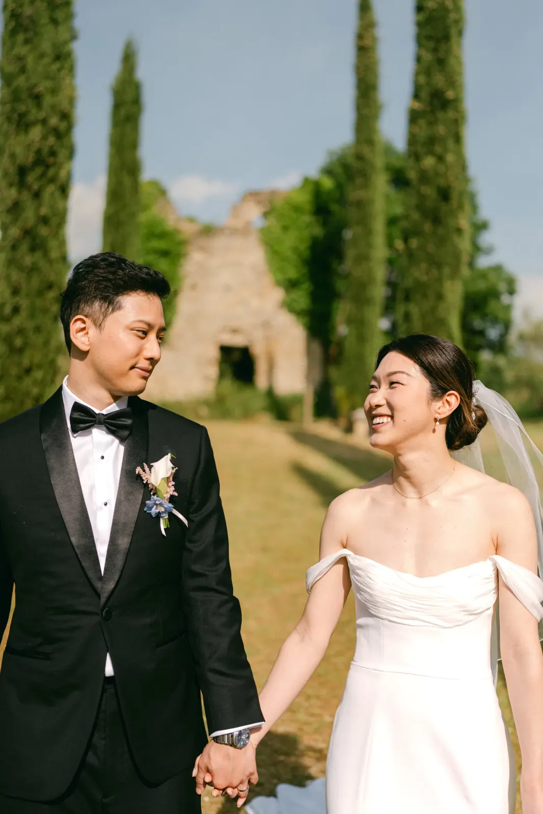 Gli sposi mano nella mano si guardano sorridendo, con i cipressi alle spalle