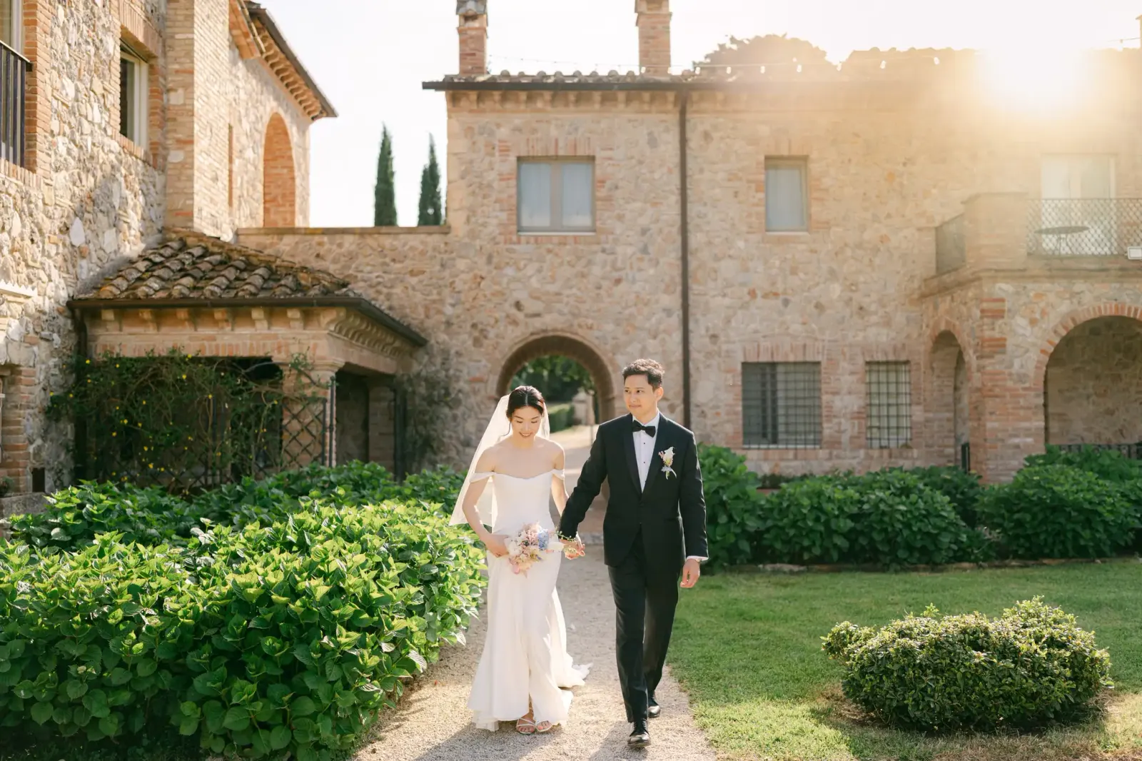 Audrey e James passeggiano in giardino con la villa in pietra alle spalle, al tramonto