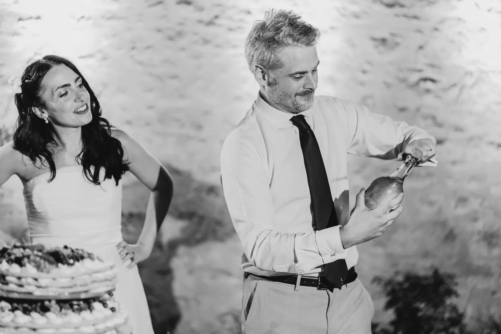 Thomas opening the sparkling wine beside the cake as Valentina watches, in black and white